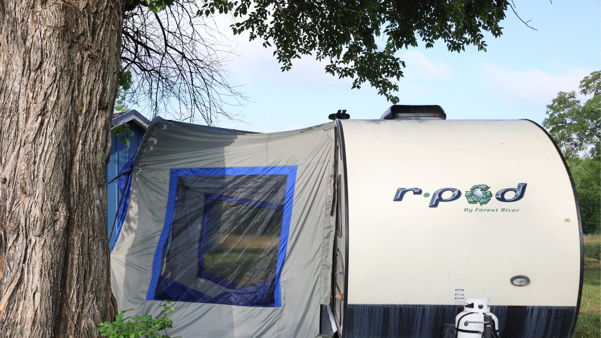 A Forest River R-Pod Trailer Side Tent by PahaQue for Enhanced Camping Comfort is pitched under a large tree, showcasing its blue-trimmed window and easy setup with camping gear—perfect for outdoor adventures. Brand: Paha Que Custom.