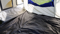 The photo shows the inside corner of the Forest River R-Pod Trailer Side Tent by Paha Que Custom, featuring a black floor, beige and white fabric walls, blue-topped window, and sunlight shining in—ideal for enhanced camping comfort.
