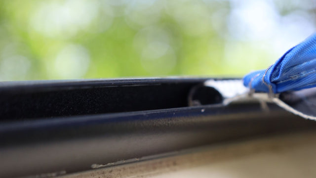 A gloved hand uses a small brush to clean dirt from the narrow groove of a Forest River R-Pod Trailer Side Tent by PahaQue, with a blurred green background highlighting this Paha Que Custom camping accessory.