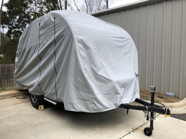 A Little Guy Mini Max FX trailer is parked on a driveway, protected by a gray PahaQue Wilderness cover made from durable MagnaTuff fabric. The locked hitch and wheels are visible near the metal building.