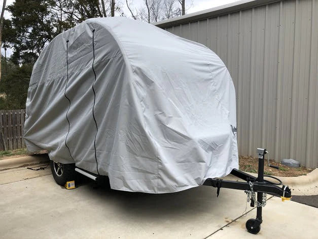 A compact camper with a Paha Que Custom NuCamp TAB 360 CS Trailer Cover by PahaQue, designed for heavy-duty, weatherproof protection, is parked on a concrete driveway by a metal building, with wheel chocks securing its tires.