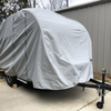A Little Guy Mini Max FX trailer is parked on a driveway, protected by a gray PahaQue Wilderness cover made from durable MagnaTuff fabric. The locked hitch and wheels are visible near the metal building.