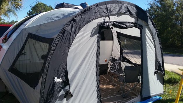 A silver Little Guy Mini Max Side Tent with black trim by PahaQue Wilderness is attached to a camper. Mesh windows and an open door reveal a folding chair inside, set up near a road on a sunny day.