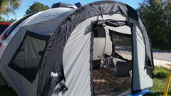 A silver Little Guy Mini Max Side Tent with black trim by PahaQue Wilderness is attached to a camper. Mesh windows and an open door reveal a folding chair inside, set up near a road on a sunny day.