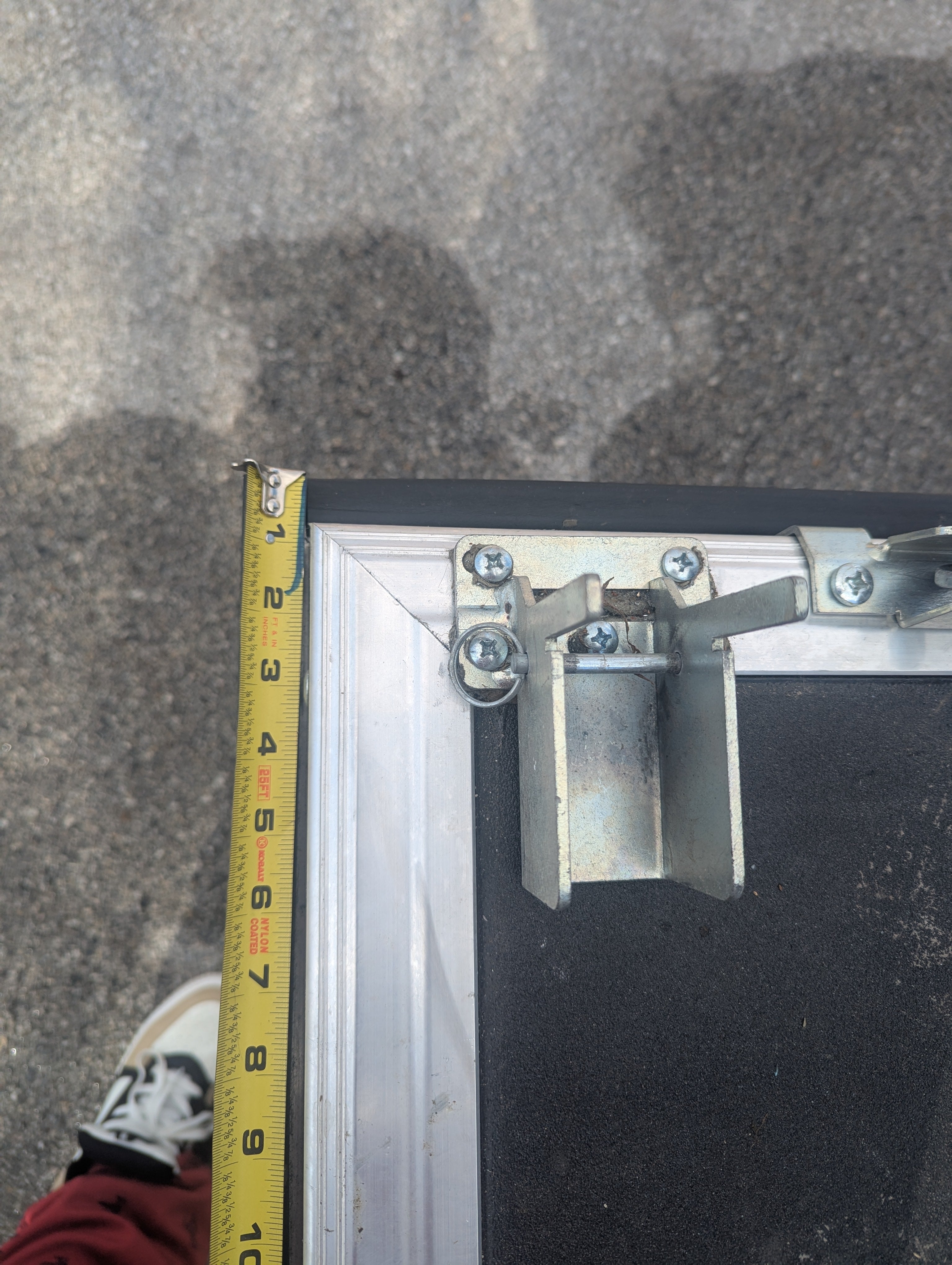 A yellow tape measure extends about 6 inches from a black-and-metal case, with a sneaker and pavement in view—suggesting setup measurements for the Custom Toy Hauler Patio Ramp Tent by PahaQue Wilderness for outdoor adventures.