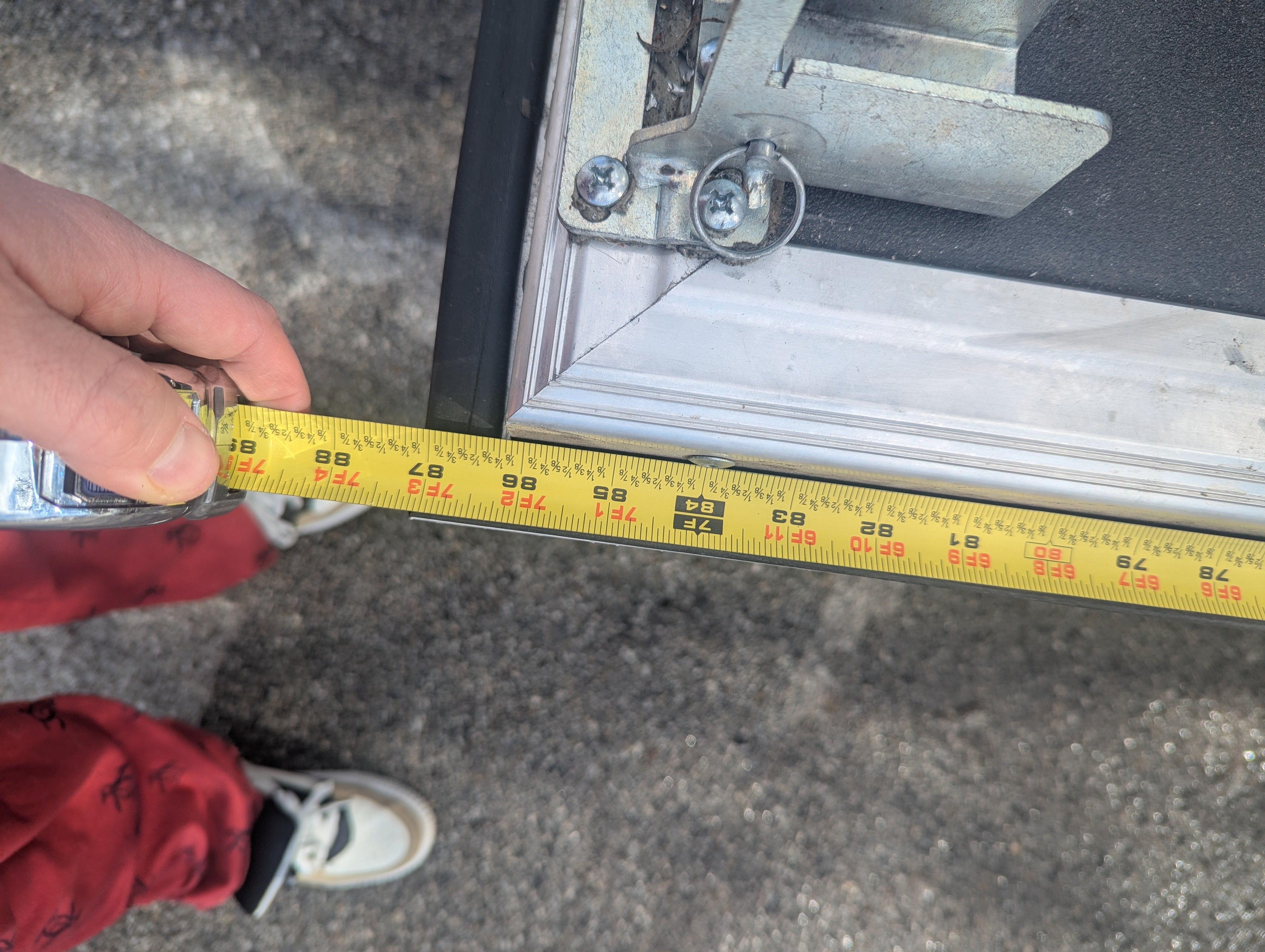 A hand measures about 49 inches across the corner of a metal-framed glass door—demonstrating how to fit the PahaQue Wilderness Custom Toy Hauler Patio Ramp Tent for outdoor adventures. The person wears red pants and white shoes on an outdoor surface.