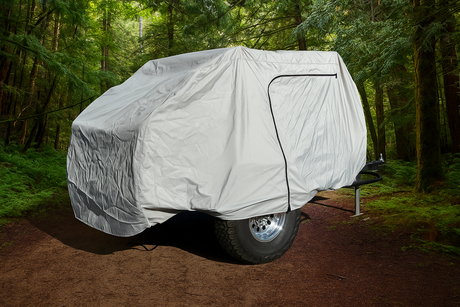 A small trailer covered by a Timberleaf Trailer Cover from Paha Que Custom sits on a dirt path in a dense, green forest. Tall trees and lush foliage surround it, creating a peaceful outdoor atmosphere.