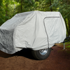 A small trailer covered by a Timberleaf Trailer Cover from Paha Que Custom sits on a dirt path in a dense, green forest. Tall trees and lush foliage surround it, creating a peaceful outdoor atmosphere.