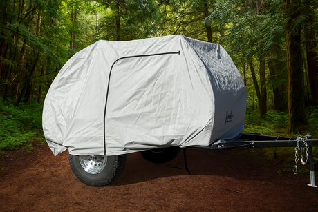 A Timberleaf teardrop trailer is parked on a forest path, shielded by a fitted gray Paha Que Custom Timberleaf Trailer Cover that protects the trailer’s large wheels and black front hitch from the elements.