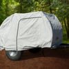 A Timberleaf teardrop trailer is parked on a forest path, shielded by a fitted gray Paha Que Custom Timberleaf Trailer Cover that protects the trailer’s large wheels and black front hitch from the elements.