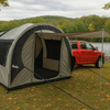 A silver and black PahaQue Wilderness Little Guy Mini Max Side Tent is set up by a lake, attached to a red pickup truck. Trees with green and yellow leaves surround the site, with hills in the background.