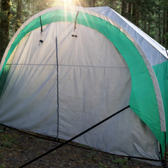 The Paha Que Custom NuCamp TAB 320 Awning Solid Front Wall Accessory for T@B 320 Trailer is set up in a sunlit forest, with sunlight streaming through the trees and illuminating the tent’s entrance.