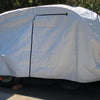 A So-Cal Teardrops Sierra Trailer is protected by a fitted gray So-Cal Teardrops Sierra Trailer Cover from PahaQue Wilderness, made of durable MagnaTuff fabric with a black center zipper, positioned on a driveway near a wooden fence and trees.