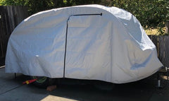 A So-Cal Teardrops Sierra Trailer is protected by a fitted gray So-Cal Teardrops Sierra Trailer Cover from PahaQue Wilderness, made of durable MagnaTuff fabric with a black center zipper, positioned on a driveway near a wooden fence and trees.