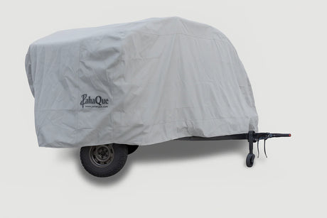 A trailer protected by a Teardrops Northwest Trailer Cover from PahaQue Wilderness, featuring a large fitted gray design with the PahaQue logo. The wheels and part of the hitch are partially visible beneath this custom-fit cover.