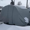 A vehicle fitted with the Ultimate Toys Trailer Cover by PahaQue from Paha Que Custom stands in a snowy lot, snow gathered on top and around it. Winter trees and a utility pole appear in the background.