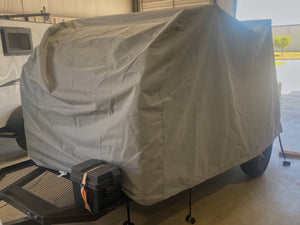 A UKAN Camp 590G/585G 5x9 Cover by PahaQue Wilderness, made of durable MagnaTuff fabric, protects a covered Little Guy Max trailer with a black storage box in a garage near a white vehicle, sunlight outside.
