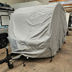 A travel trailer is parked indoors on concrete, protected by the Ultimate Toys Trailer Cover By PahaQue Durable Weather-Resistant Protection from Paha Que Custom, with other RVs and camping vehicles visible in the background.