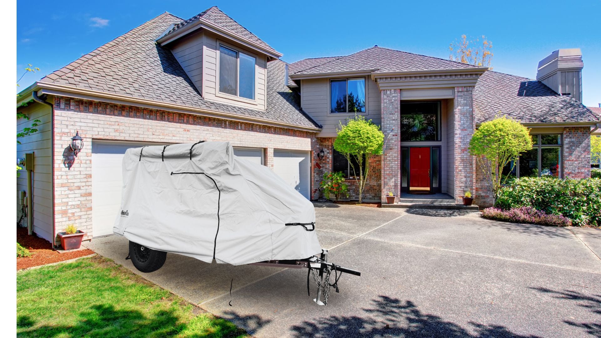 A trailer protected by the PahaQue Wilderness Zion Off Road Trailer Cover, made from heavy-duty MagnaTuff fabric, sits on the driveway before a large brick house with a red door, attached garage, and neat bushes.
