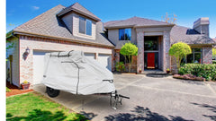 A trailer protected by the PahaQue Wilderness Zion Off Road Trailer Cover, made from heavy-duty MagnaTuff fabric, sits on the driveway before a large brick house with a red door, attached garage, and neat bushes.