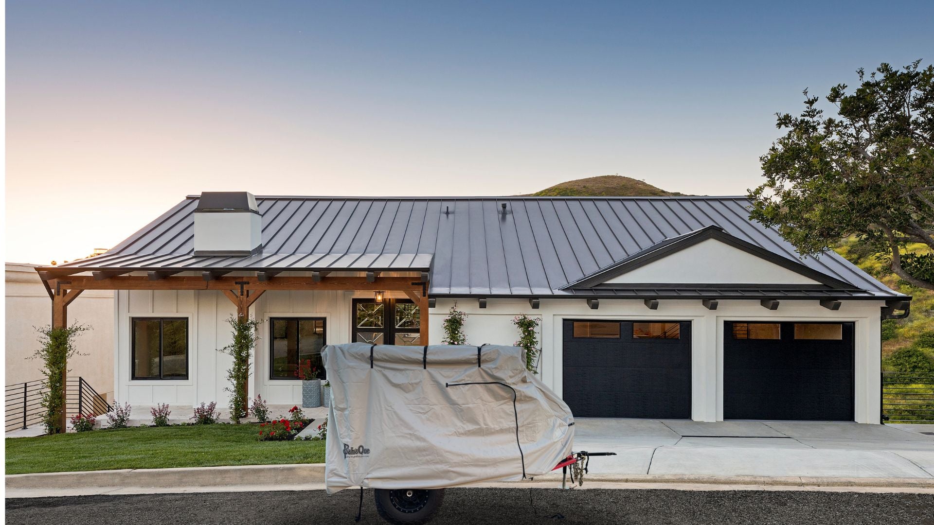 A modern house with a metal roof, white exterior, and two black garage doors has a covered trailer in the driveway featuring the Zion Off Road Trailer Cover Heavy Duty MagnaTuff Fabric by PahaQue Wilderness. Green grass, plants, a tree, and hills are in the background.