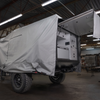 A rugged off-road trailer with large tires is partially covered by a Paha Que Custom Xpedition Trailer Cover for Ultimate Protection and Longevity, parked in a spacious industrial workshop. One end is open, showing storage compartments and protected interiors.