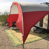 A NuCamp TAB 400 Trailer Awning by Paha Que Custom is set up on a TAB 400 trailer at a desert campsite. Under the red and white awning are camping chairs and a woven mat, with desert plants and cacti in the background.