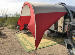 A NuCamp TAB 400 Trailer Awning by Paha Que Custom is set up on a TAB 400 trailer at a desert campsite. Under the red and white awning are camping chairs and a woven mat, with desert plants and cacti in the background.