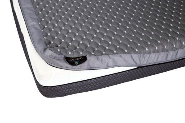 Close-up of a grey, quilted Divini Mattress topper by PahaQue Wilderness, featuring a geometric pattern and split-side firmness, on a white mattress with a dark border. A small black label shows the Divini branding.