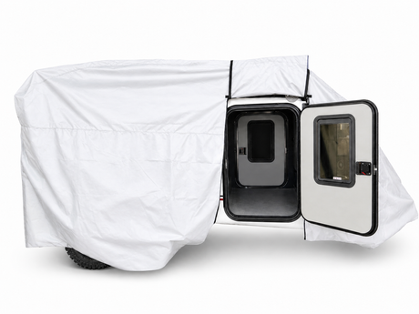 A small trailer is partially covered with the Encore Trailer Cover by Paha Que Custom, offering premium protection. The open side door reveals a dark interior with black trim, a window in the door, and visible off-road tires.