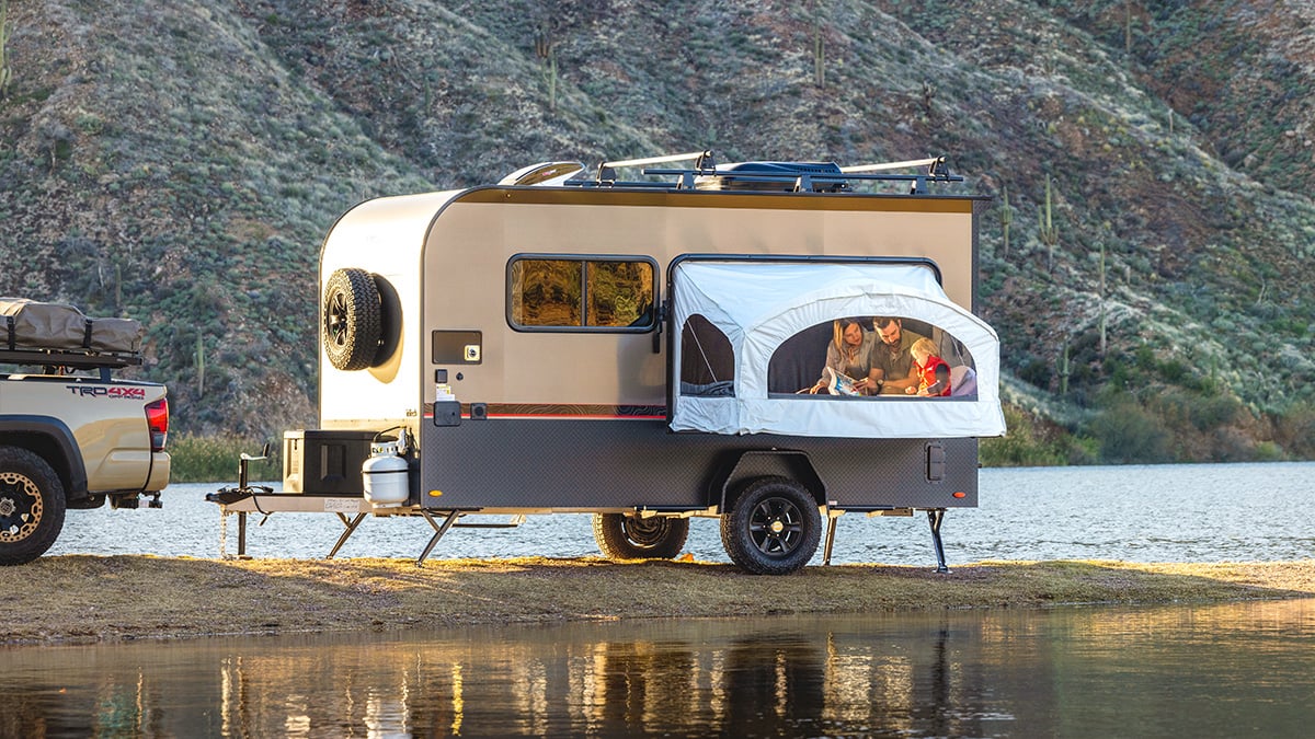 A family enjoys lake and mountain views from inside the weatherproof Toy Hauler Patio Ramp Tent by PahaQue for Intech Trailers, while their truck is parked beside the camper.
