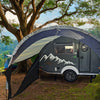 A gray camper with a mountain design sits in a grassy area beneath trees and mountains. A Paha Que Custom NuCamp TAB 320 Trailer Awning 2 by PahaQue—NEW FOR 2024—plus mesh side panels offers shade beside the trailer.