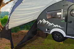 A gray teardrop camper with a mountain graphic is parked on grass, partly shaded by the NuCamp TAB 320 Trailer Awning 2 by Paha Que Custom—NEW FOR 2024. Green trees and blue sky are seen in the background.