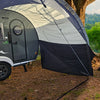 A small gray camper with a NuCamp TAB 320 Trailer Awning 2 by PahaQue, and a tan tent are set up under a large tree in a lush, green outdoor setting, partially framed by trees and grass. Brand: Paha Que Custom.