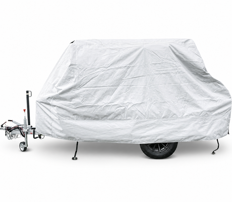 A trailer is parked on a white background, covered by the Hero Trailer Cover by PahaQue - CLEARANCE SALE. The light gray protective tarp from Paha Que Custom shields the trailer, with its wheels and towing hitch partly visible beneath.