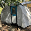 A Paha Que Custom Helio O Series Trailer sits under a tree beside a house, its blue trim and door visible beneath the Heavy Duty MagnaTuf Protection cover by PahaQue. Sunlight and shadows highlight the cover’s sturdy, weatherproof design.