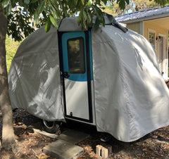 A Paha Que Custom Helio O Series Trailer sits under a tree beside a house, its blue trim and door visible beneath the Heavy Duty MagnaTuf Protection cover by PahaQue. Sunlight and shadows highlight the cover’s sturdy, weatherproof design.