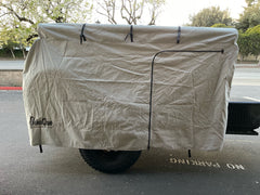 A tan PahaQue Wilderness Vintage Trailer Works XTR Cover, made from durable MagnaTuff fabric and featuring a zippered door, protects an outdoor grill or smoker mounted on a trailer with large black tires parked near a NO PARKING curb.