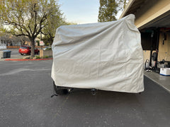 A vehicle is parked in a driveway near a garage, protected by the PahaQue Wilderness Vintage Trailer Works XTR Cover Heavy Duty Protection for Vintage Trailers. Trees and parked cars are visible in the background.