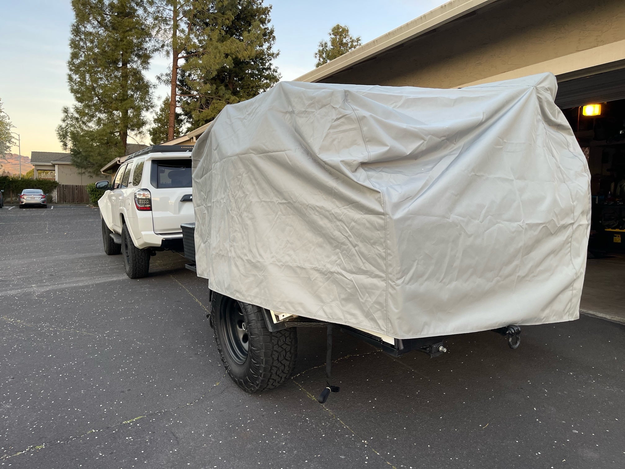 A white SUV is parked in a driveway by a building, towing a trailer covered with the PahaQue Wilderness Vintage Trailer Works XTR Cover Heavy Duty Protection for Vintage Trailers. Trees and a fence are in the background.