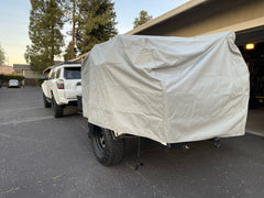 A white SUV is parked in a driveway by a building, towing a trailer covered with the PahaQue Wilderness Vintage Trailer Works XTR Cover Heavy Duty Protection for Vintage Trailers. Trees and a fence are in the background.