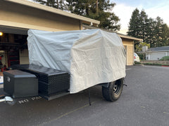 A small utility trailer is parked in a driveway, protected by the PahaQue Wilderness Vintage Trailer Works XTR Cover Heavy Duty Protection for Vintage Trailers. Trees and houses can be seen in the background on a clear day.