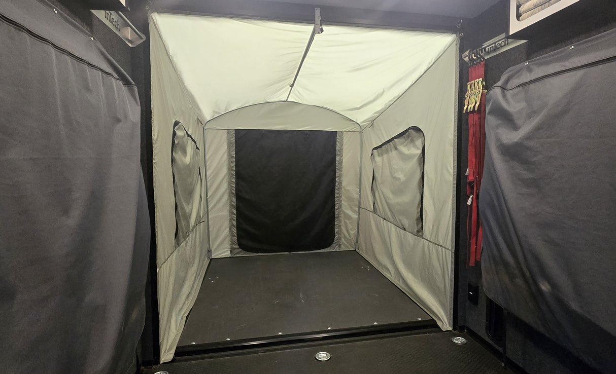 A PahaQue Wilderness Toy Hauler Patio Enclosure Ramp Tent, custom built to fit all toy hauler trailers, is attached to the back of a vehicle and features zippered windows and a door. The gray enclosure is viewed from inside the cargo area.