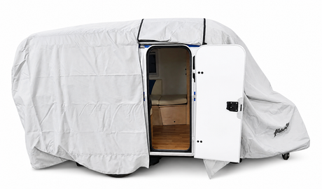 A small camper trailer fitted with the Paha Que Custom Meerkat Trailer Cover, made from heavy-duty MagnaTuff fabric for weather-resistant protection in gray, sits with its side door open to show a wooden floor and beige seating inside.