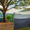 A campsite features a white vehicle with a NuCamp TAB 320 Trailer Awning 2 by PahaQue (NEW FOR 2024) from Paha Que Custom, set up with mesh side panels. Nearby, a blue camping chair and table overlook green hills on a sunny day.