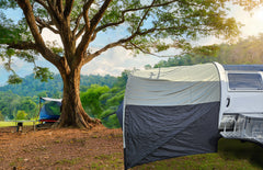A campsite features a white vehicle with a NuCamp TAB 320 Trailer Awning 2 by PahaQue (NEW FOR 2024) from Paha Que Custom, set up with mesh side panels. Nearby, a blue camping chair and table overlook green hills on a sunny day.