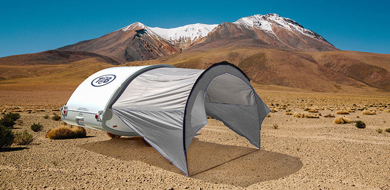 A small white teardrop camper with a Paha Que Custom NuCamp TAB 320 Trailer Awning is set up on a dry, rocky desert plain, its gray tent providing shade as tall mountains rise under a clear blue sky.