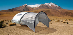 A small white teardrop camper with a Paha Que Custom NuCamp TAB 320 Trailer Awning is set up on a dry, rocky desert plain, its gray tent providing shade as tall mountains rise under a clear blue sky.