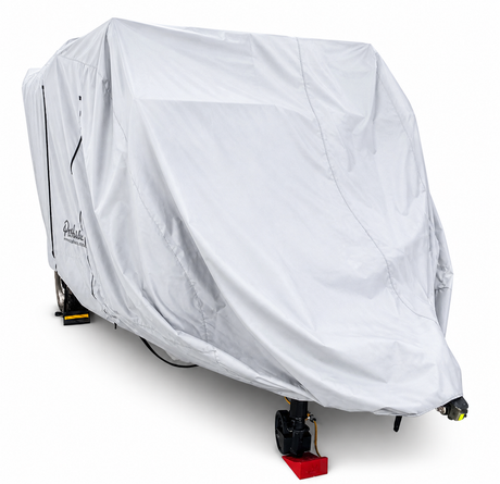 A Teton X travel trailer is fully enclosed in the Paha Que Custom Teton X Trailer Cover Premium Protection, concealing its features for superior weather protection, with only the wheels and hitch partially visible on a plain white background.