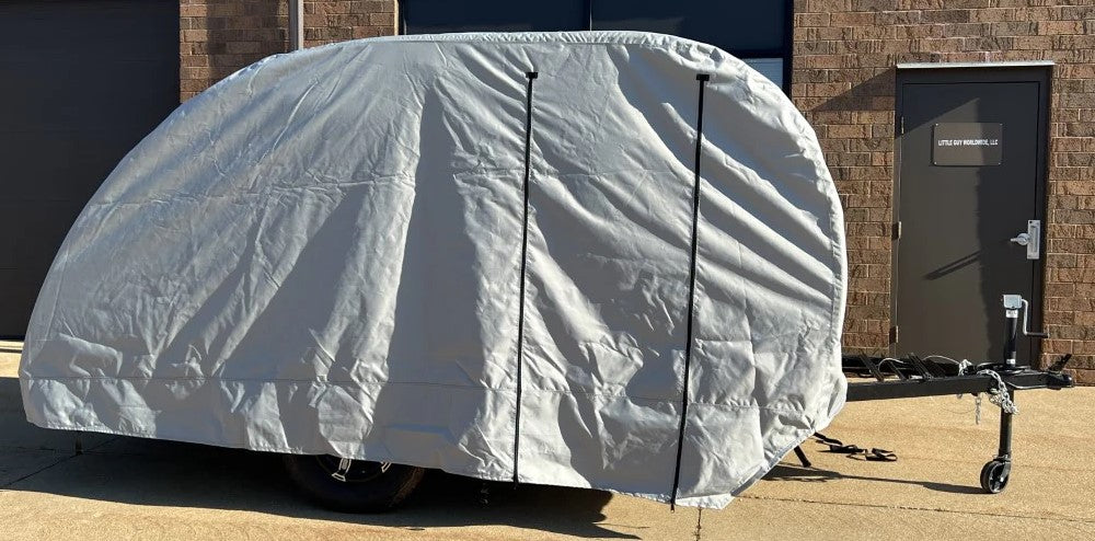 A Little Guy Micro Max trailer is parked on concrete outside a brick building, fully wrapped in a PahaQue Wilderness Little Guy Micro Max Cover, made from durable MagnaTuff fabric with only the hitch and wheels partially visible.